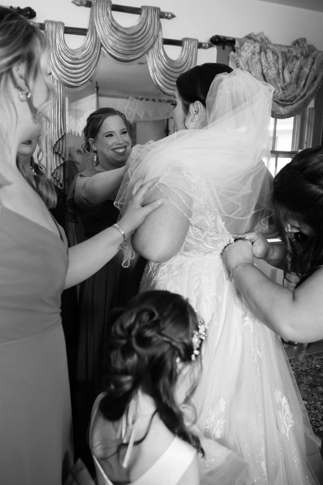 Black and white photo of bridesmaids helping a bride into her lace wedding dress before the ceremony.