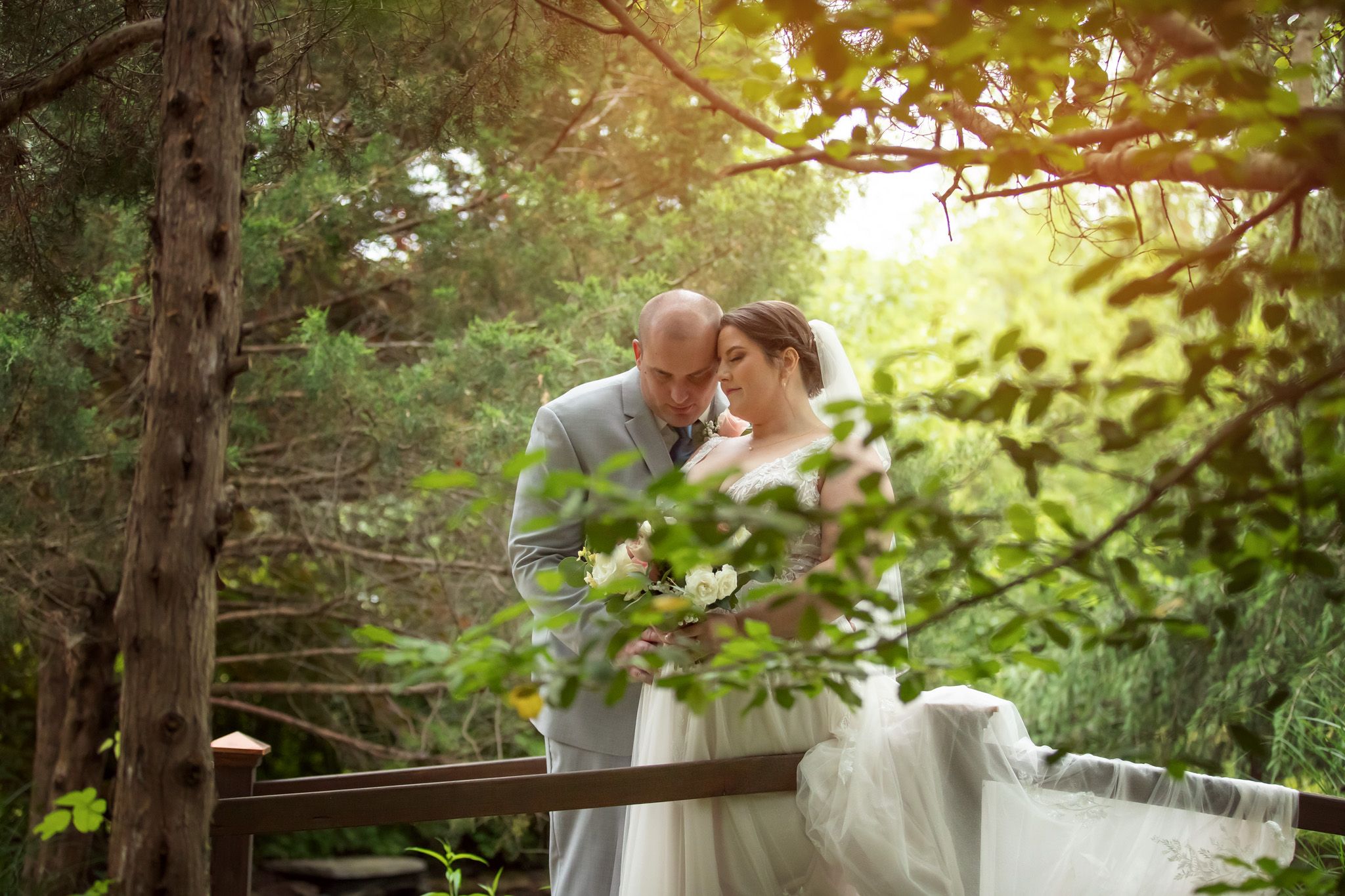 A tender moment between a bride and groom on a wooden bridge surrounded by lush green forest trees.