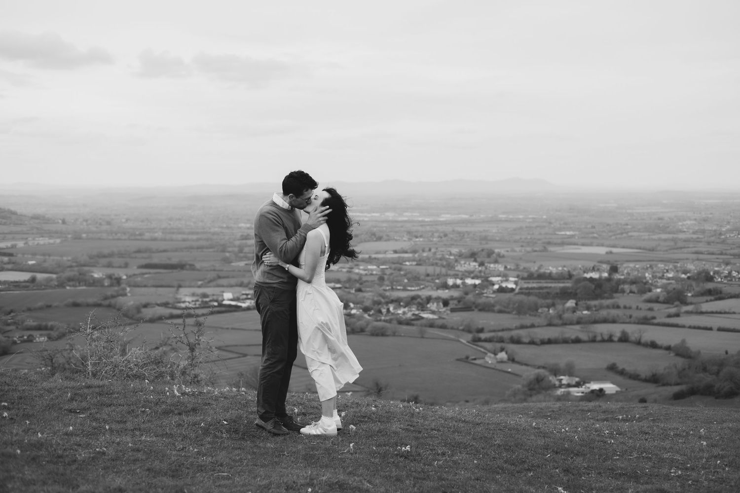 Black and white photo of a couple kissing on a hilltop with scenic countryside views stretching into the distance.