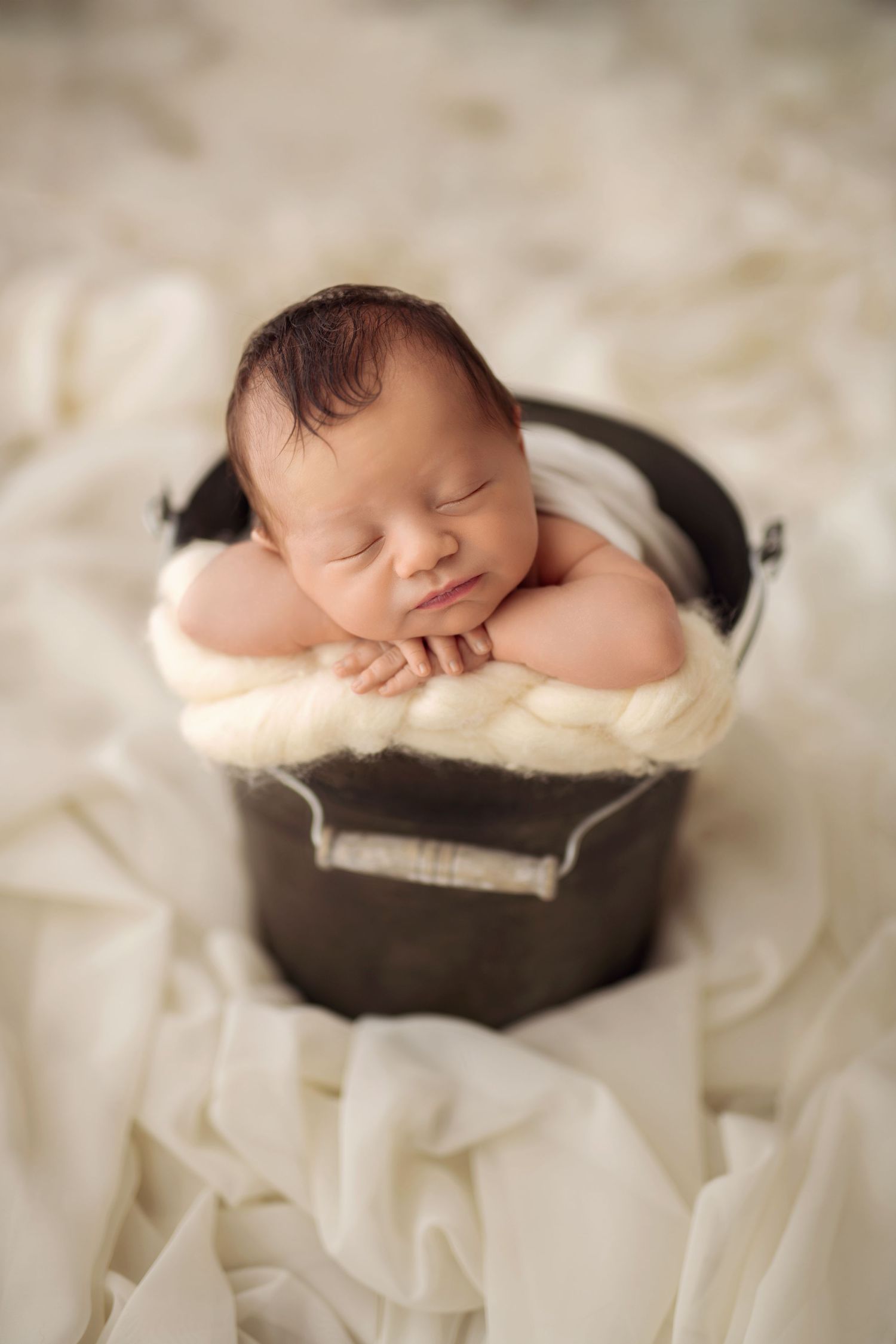 Sleeping newborn baby with dark hair resting chin on hands inside a small black bucket prop.