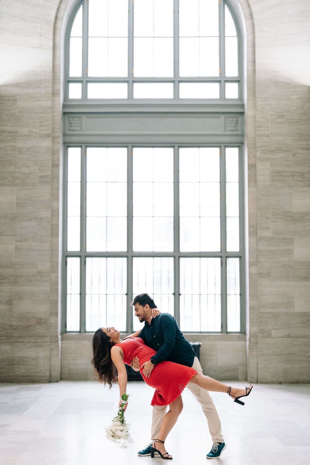 Couple dancing romantically in elegant hall, woman in red dress being dipped by man in dark jacket near large arched windows.