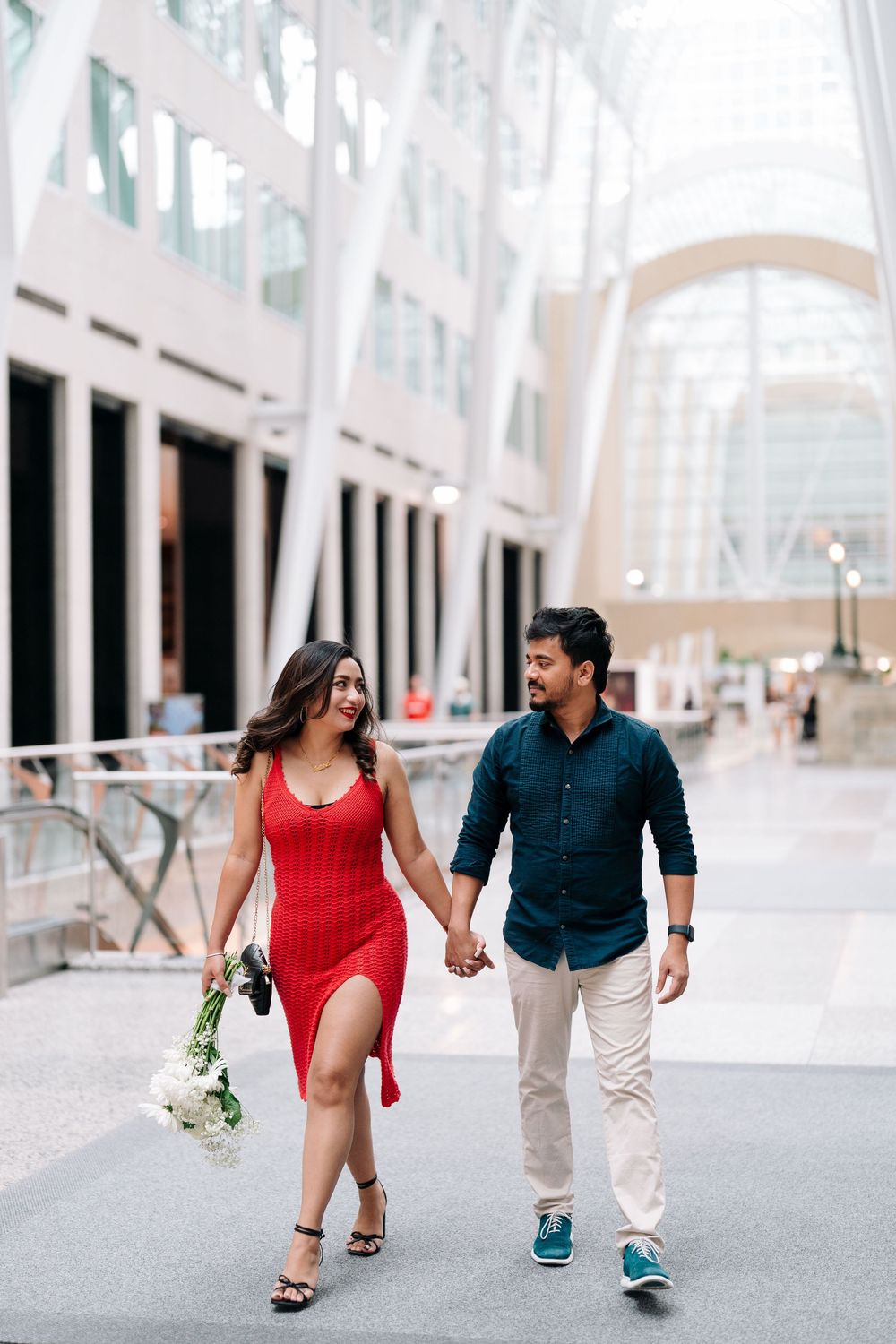 Couple holding hands walking through modern atrium, woman in red dress with flowers, man in navy shirt and khakis.