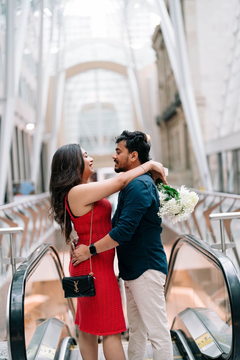 Romantic couple embracing on escalator, woman in red dress holding white flowers in modern shopping mall.