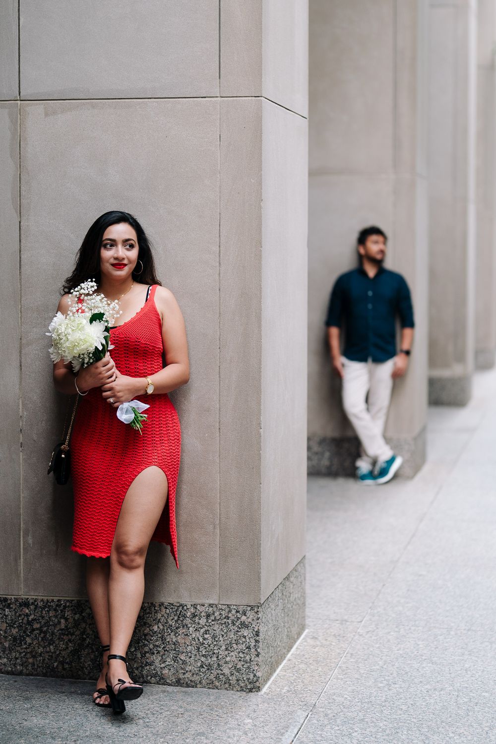 Woman in red dress holding white flowers leans against stone column, man in blue shirt stands blurred in background.