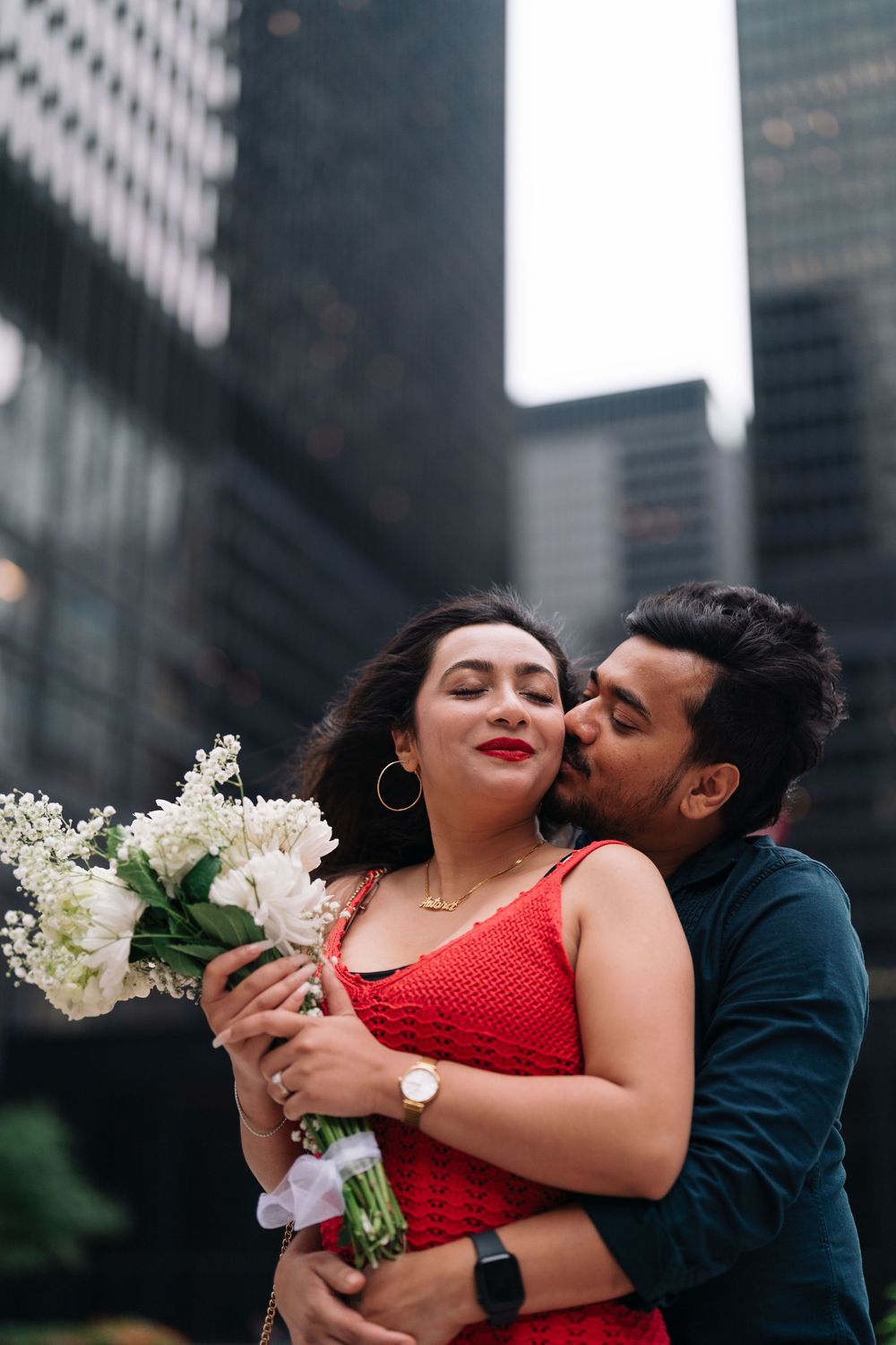 Romantic couple embracing in city, woman in red dress holding white flowers, man kissing her cheek near skyscrapers.