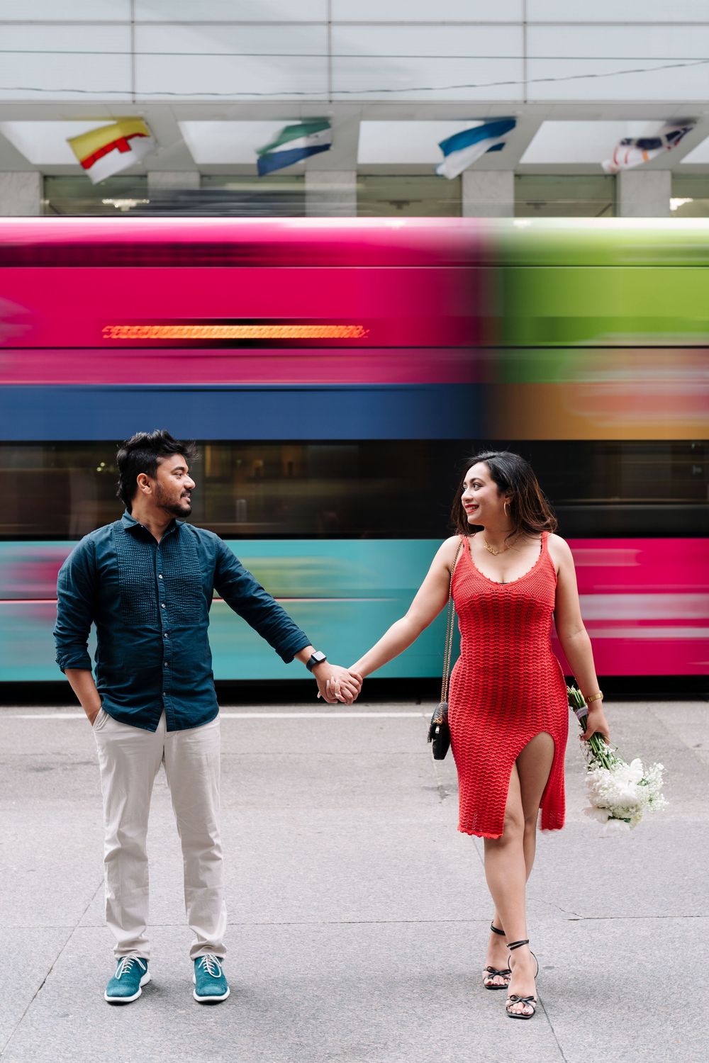 Couple holding hands on city sidewalk, woman in red dress with flowers, colorful bus passing in background.