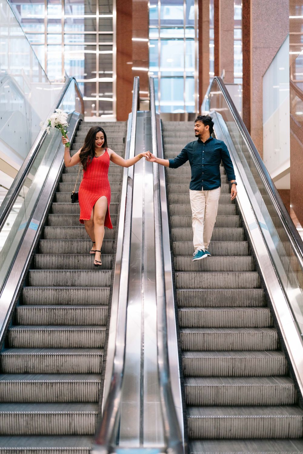 Couple holding hands on escalator, woman in red dress with bouquet, man in dark shirt and khaki pants.