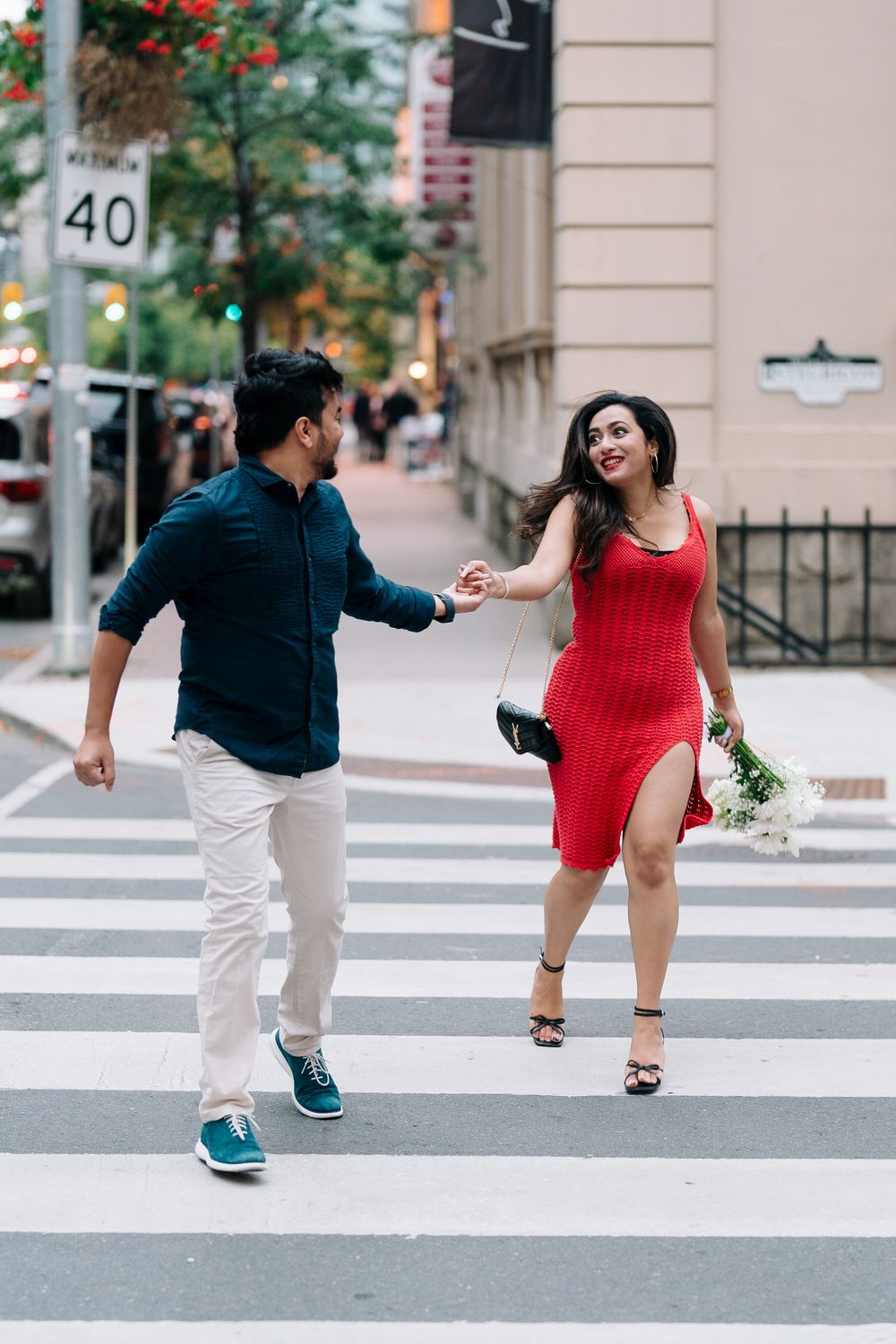 Couple crossing a city street, woman in red dress holding flowers, man in teal shirt, urban crosswalk scene.