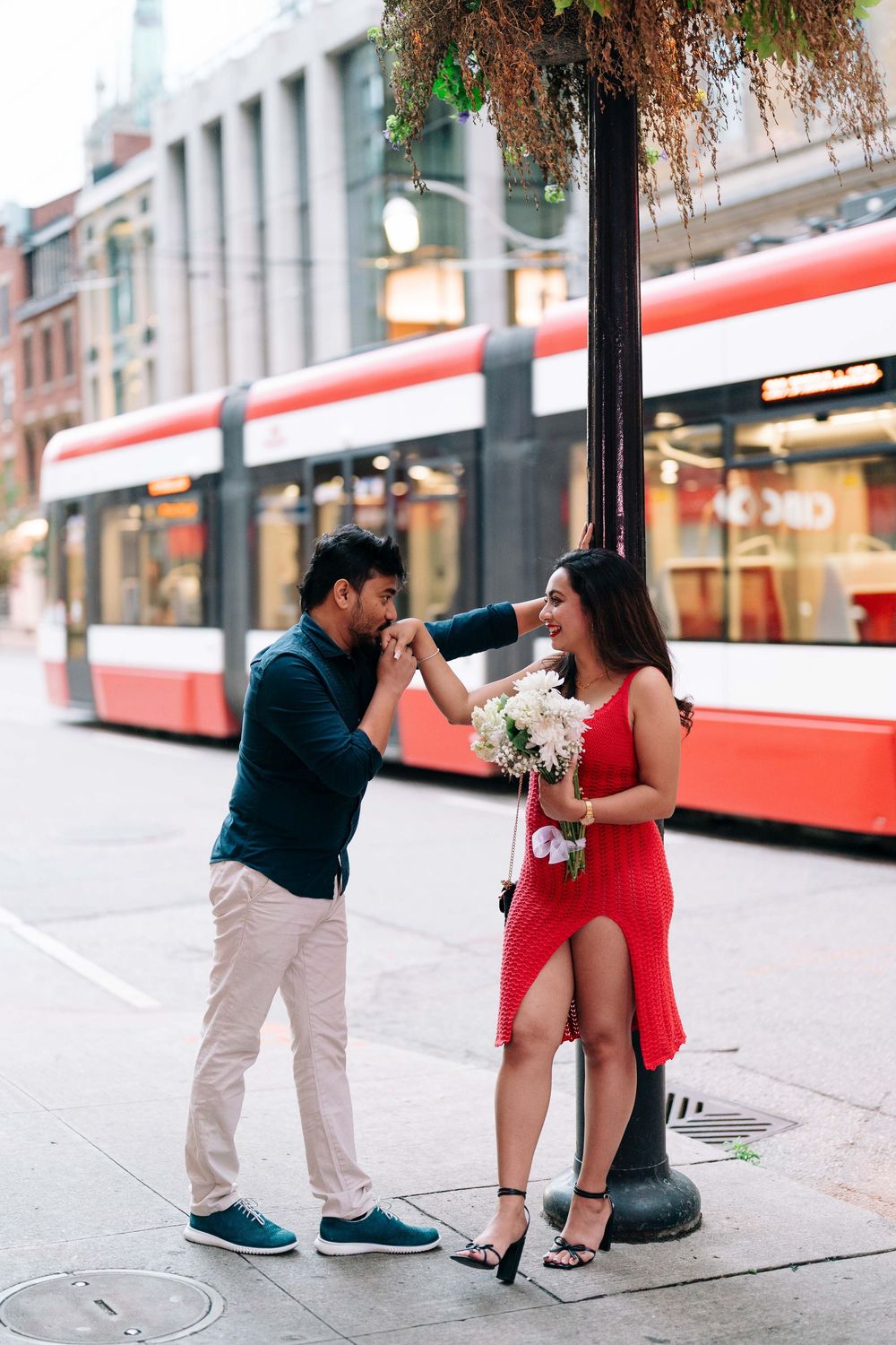 Romantic couple on city street, man kissing woman's hand, she holds white bouquet in red dress, red tram behind.