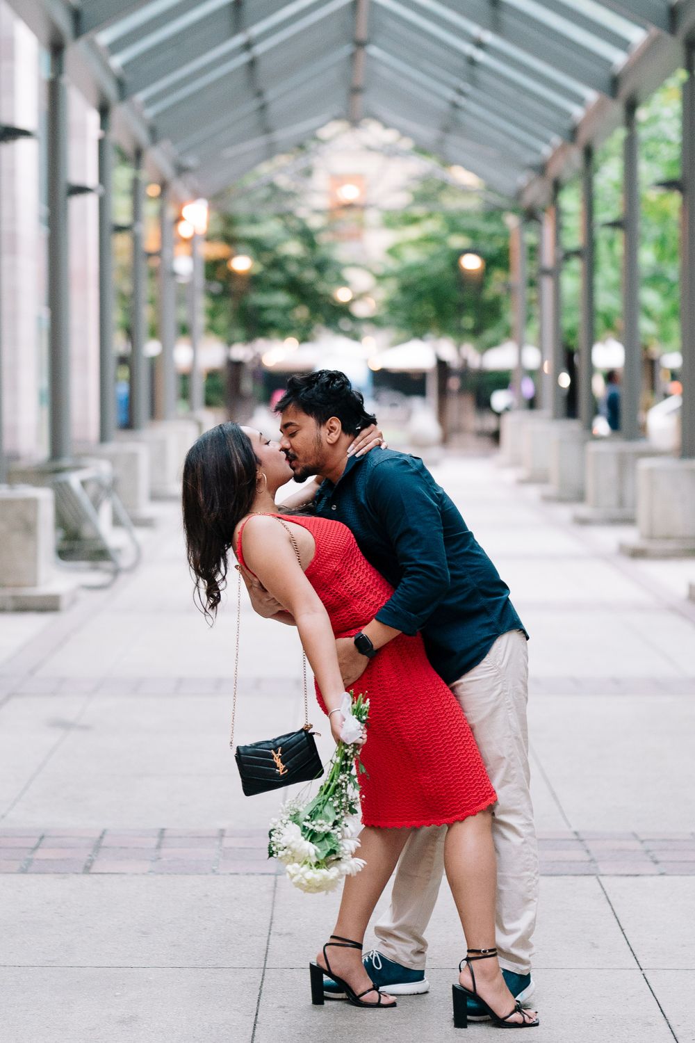 Couple sharing a romantic kiss under an urban pergola, woman in red dress holding white flowers, city backdrop.