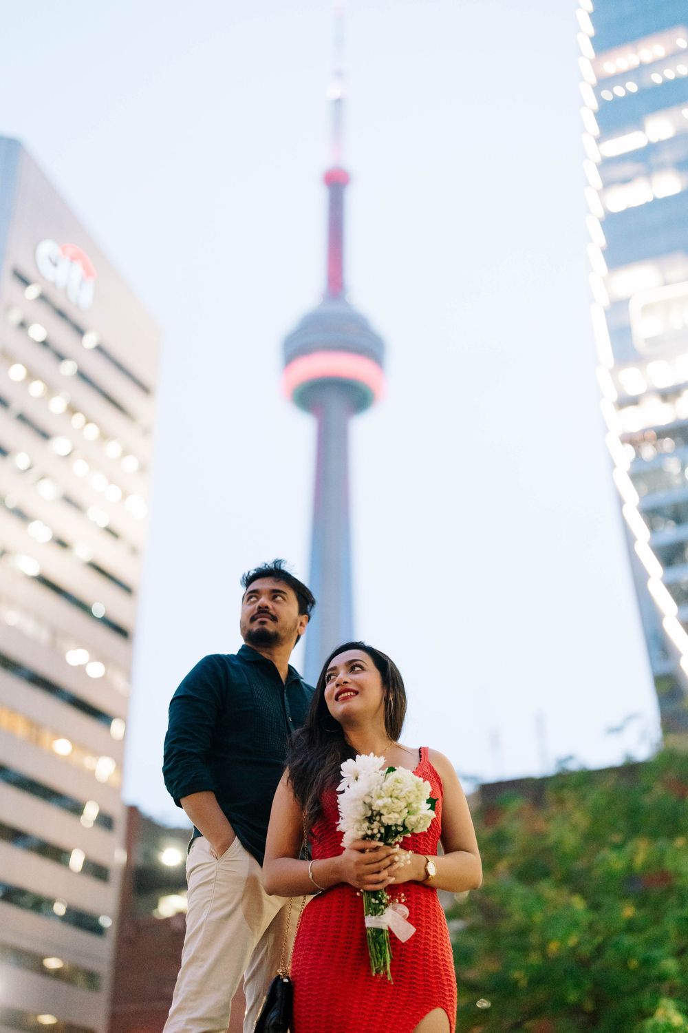 Couple posing in front of CN Tower in Toronto, woman in red dress holding white flowers bouquet at dusk.