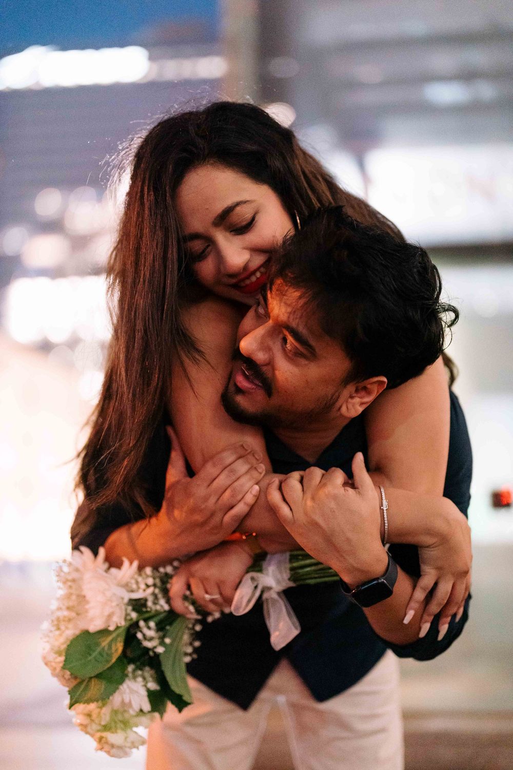 A couple shares a romantic embrace, the woman hugging the man from behind while holding a bouquet of white flowers.