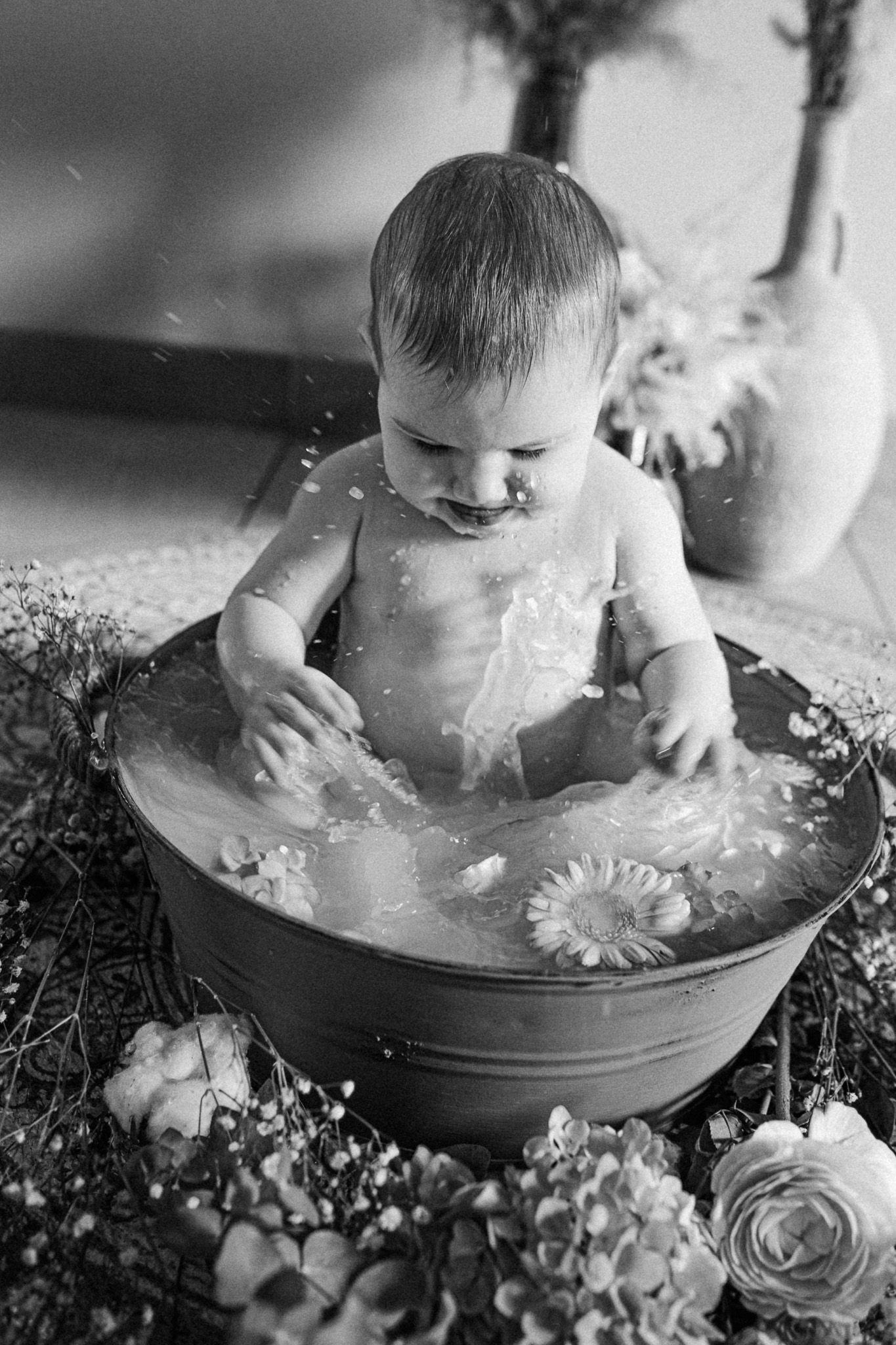 Photo en noir et blanc d’un bébé éclaboussant dans un bain fleuri (type bain de lait), entouré de fleurs.
