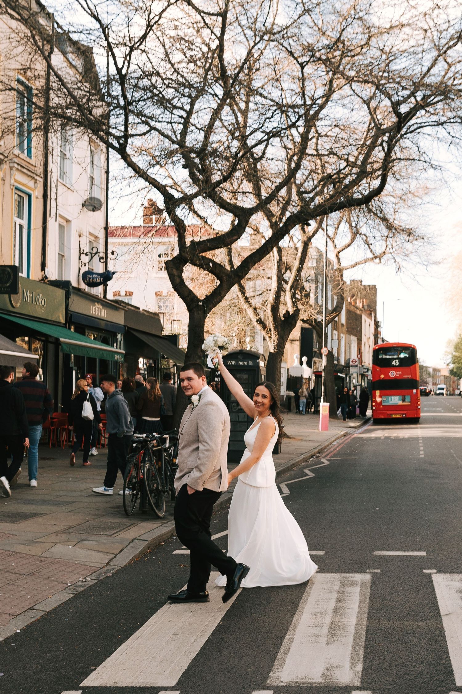 Bride and groom crossing a zebra crossing on a busy London street with a red double-decker bus in the background.