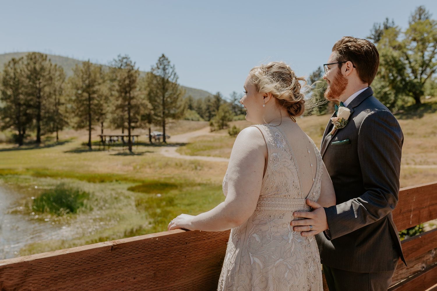 Bride and groom stand on a wooden bridge overlooking a scenic mountain pond and pine trees on their wedding day at Lake Cuyamaca, Julian.