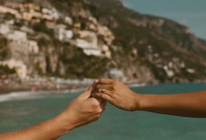 Two hands touching with a ring near the Amalfi Coast, Italy, with turquoise water and cliffside buildings in background.