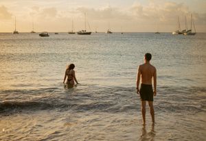Couple wading in calm tropical ocean waters at sunset with sailboats anchored in the background.