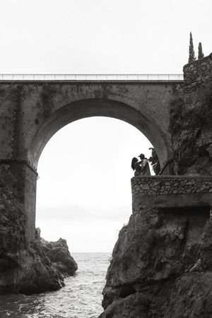 Black and white photo of a couple standing on rocky cliffs beneath a stone arch bridge overlooking the sea during their destination wedding.