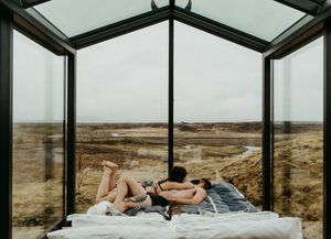 Couple relaxing on a bed in a glass cabin with panoramic views of a vast, serene landscape under overcast skies.