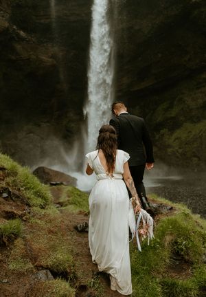 A bride in a white gown and groom in a black suit walk toward a stunning waterfall surrounded by lush greenery.