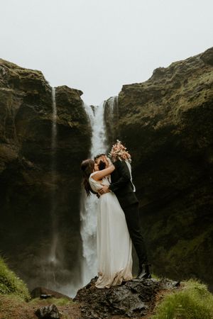 Couple kissing in front of a dramatic waterfall between mossy cliffs during their outdoor elopement ceremony.