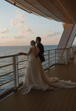 Bride and groom stand together on cruise ship deck at sunset, overlooking the ocean, wedding dress train flowing behind.