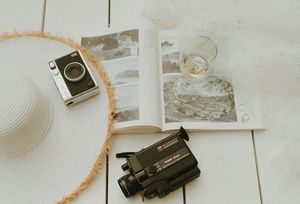 Vintage camera, sun hat, open magazine, glass of white wine, and retro camcorder arranged on white wooden surface.