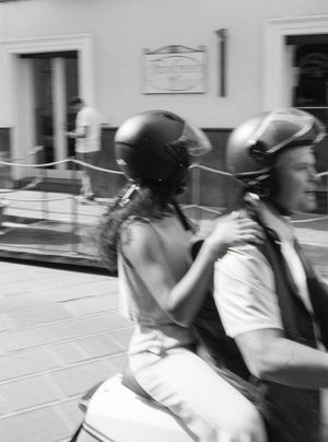 Black and white photo of two people riding a scooter wearing helmets on a city street.