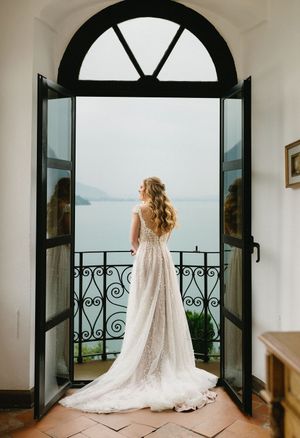 Bride in elegant lace wedding gown stands at arched French doors overlooking a scenic lake and mountains.