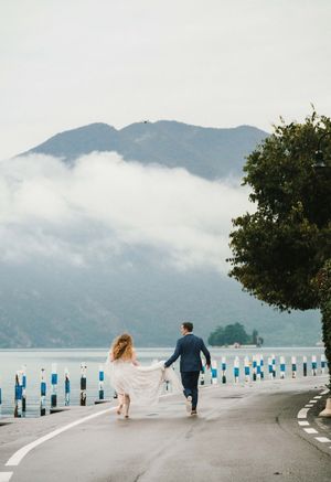 Bride and groom holding hands running along a scenic lakeside road with misty mountains in the background.