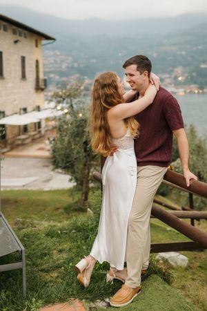 Couple embracing and sharing a romantic moment on a terrace overlooking a scenic lake and village in Italy.