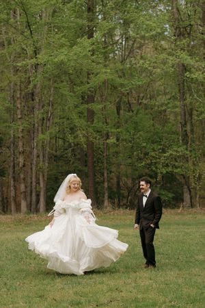 Bride twirls in white gown as groom smiles in black tuxedo in a green forest meadow.