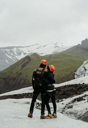 Couple in orange helmets and hiking gear embrace on a glacier with snow-capped mountains behind them.