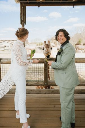 Same-sex couple in wedding attire feeding a giraffe at a zoo, bride in white lace jumpsuit and spouse in sage green suit.
