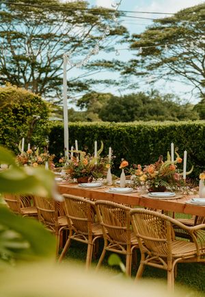Elegant outdoor wedding reception with rattan chairs, wooden table, colorful floral centerpieces, and candles set in a lush garden.