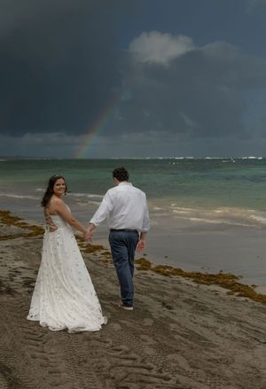 Bride and groom hold hands on a stormy beach with a rainbow over the ocean.