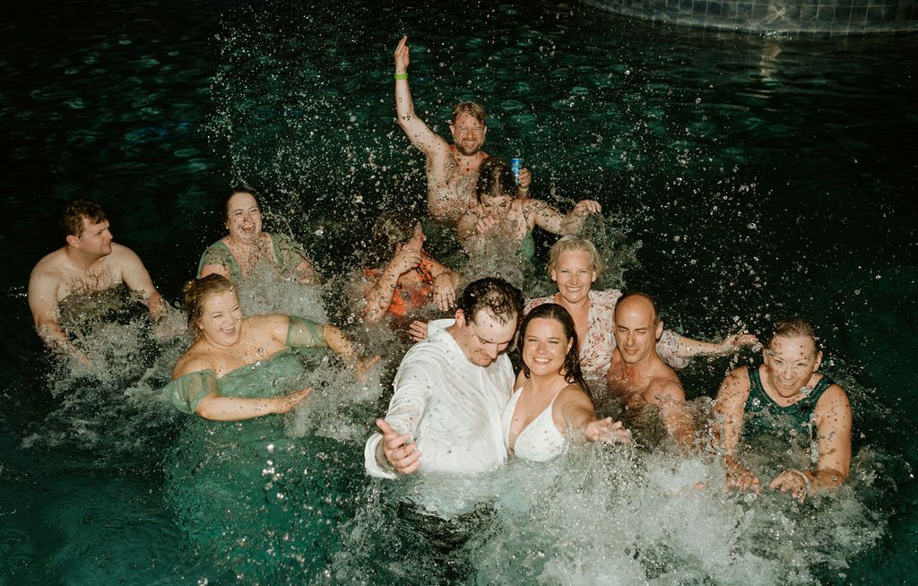 Wedding party joyfully splashing in a pool at night, bride and groom surrounded by celebrating guests.