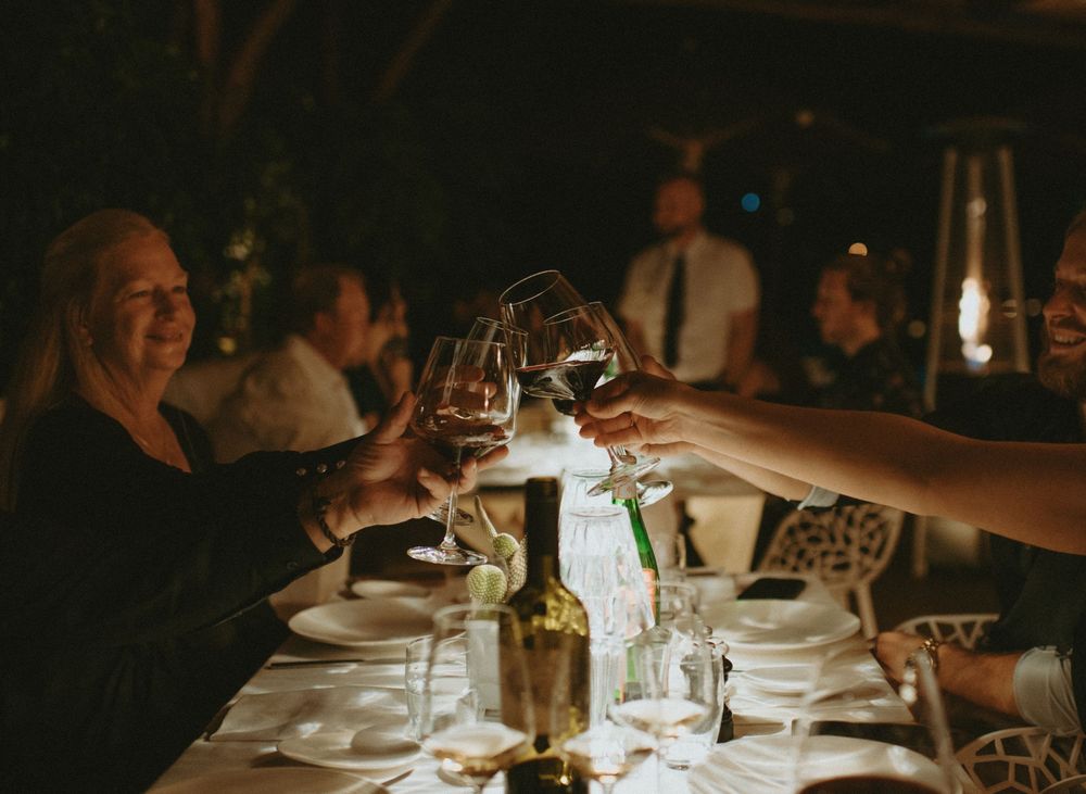 Guests raising glasses of red wine in a toast at an elegant destination wedding dinner party at night.