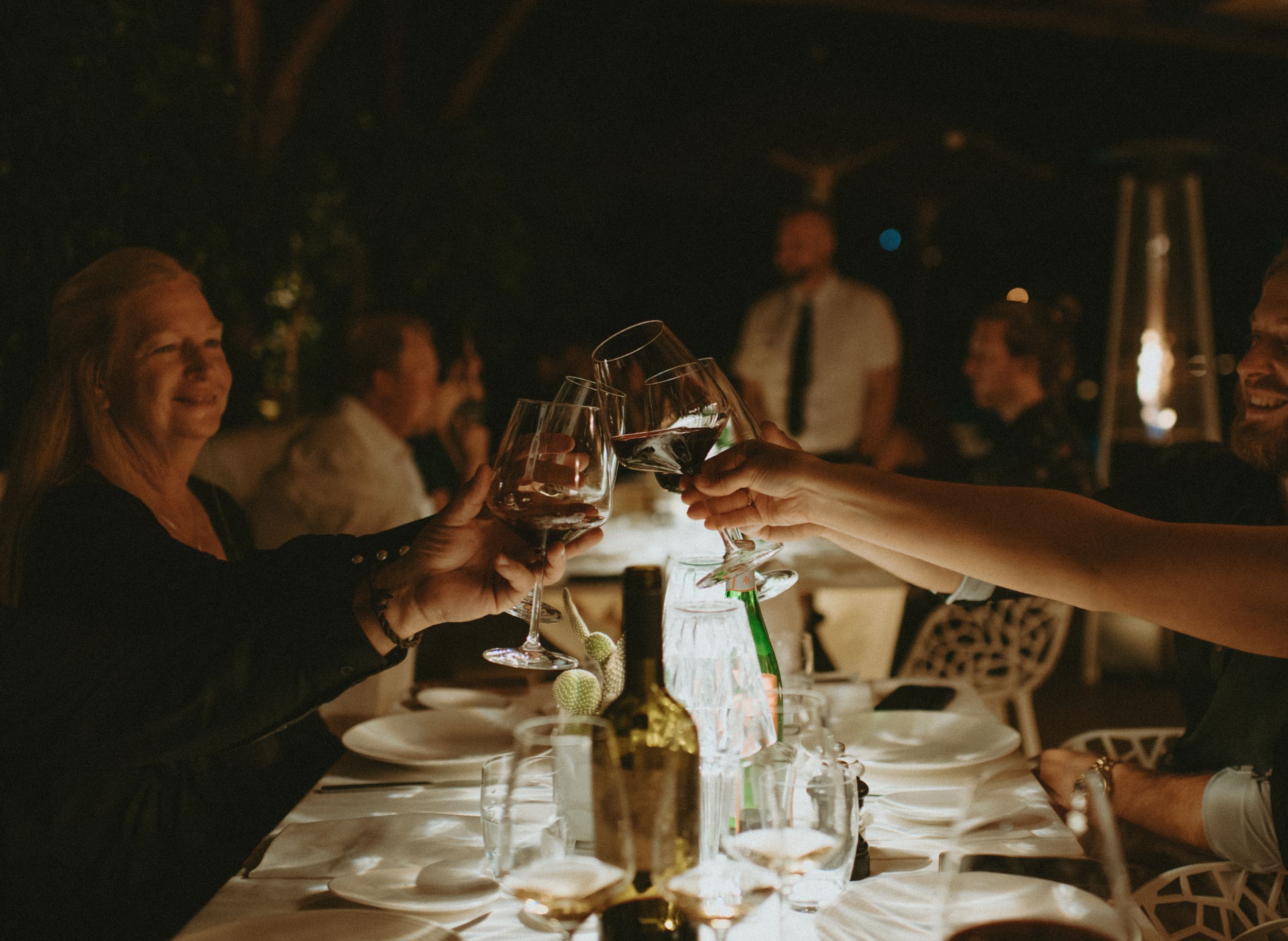 Guests raising glasses of red wine in a toast at an elegant destination wedding dinner party at night.