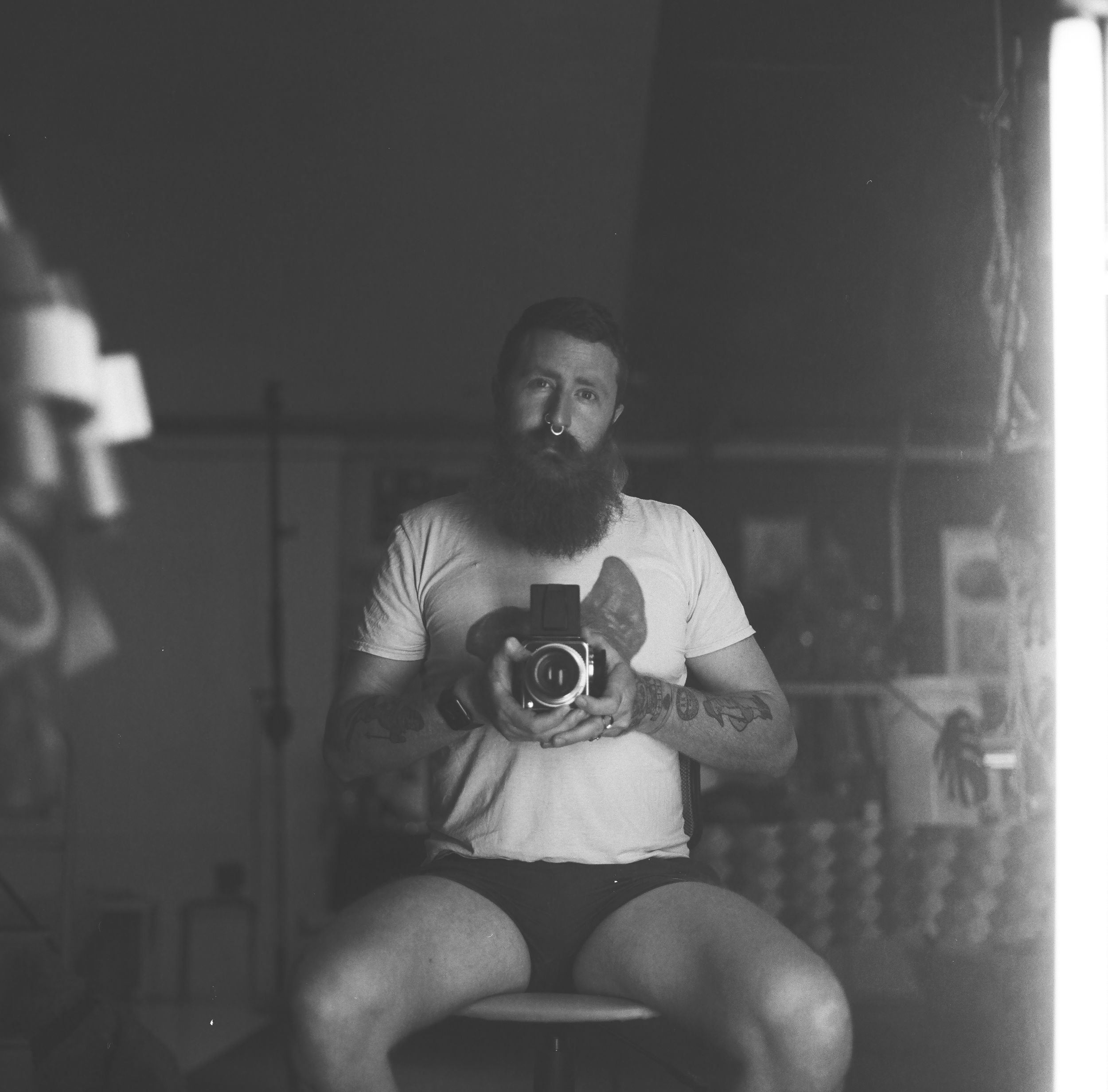 Bearded man with tattoos takes a mirror selfie holding a vintage camera, wearing a white t-shirt, black and white photo.