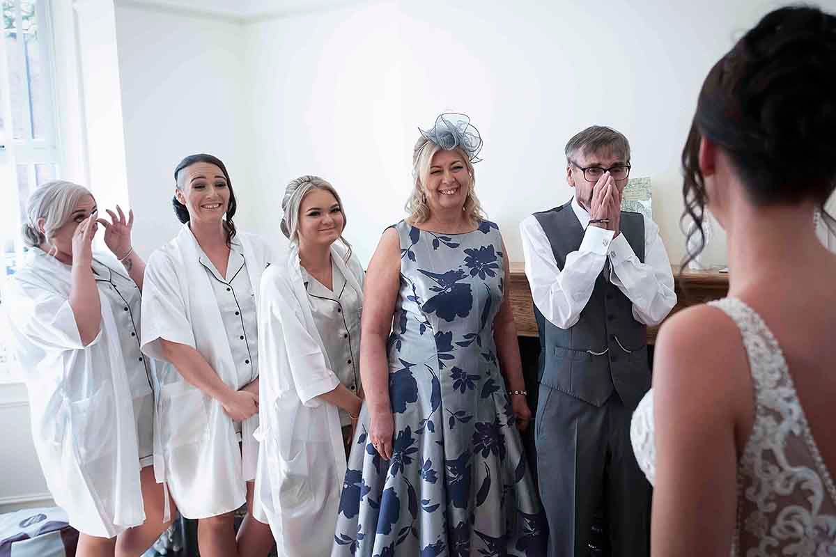 Bridesmaids in white robes and guests react with joy and tears as bride reveals her wedding dress to her father