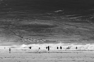Black and white photo of surfers and beachgoers on a vast sandy beach with large waves and a hillside backdrop.