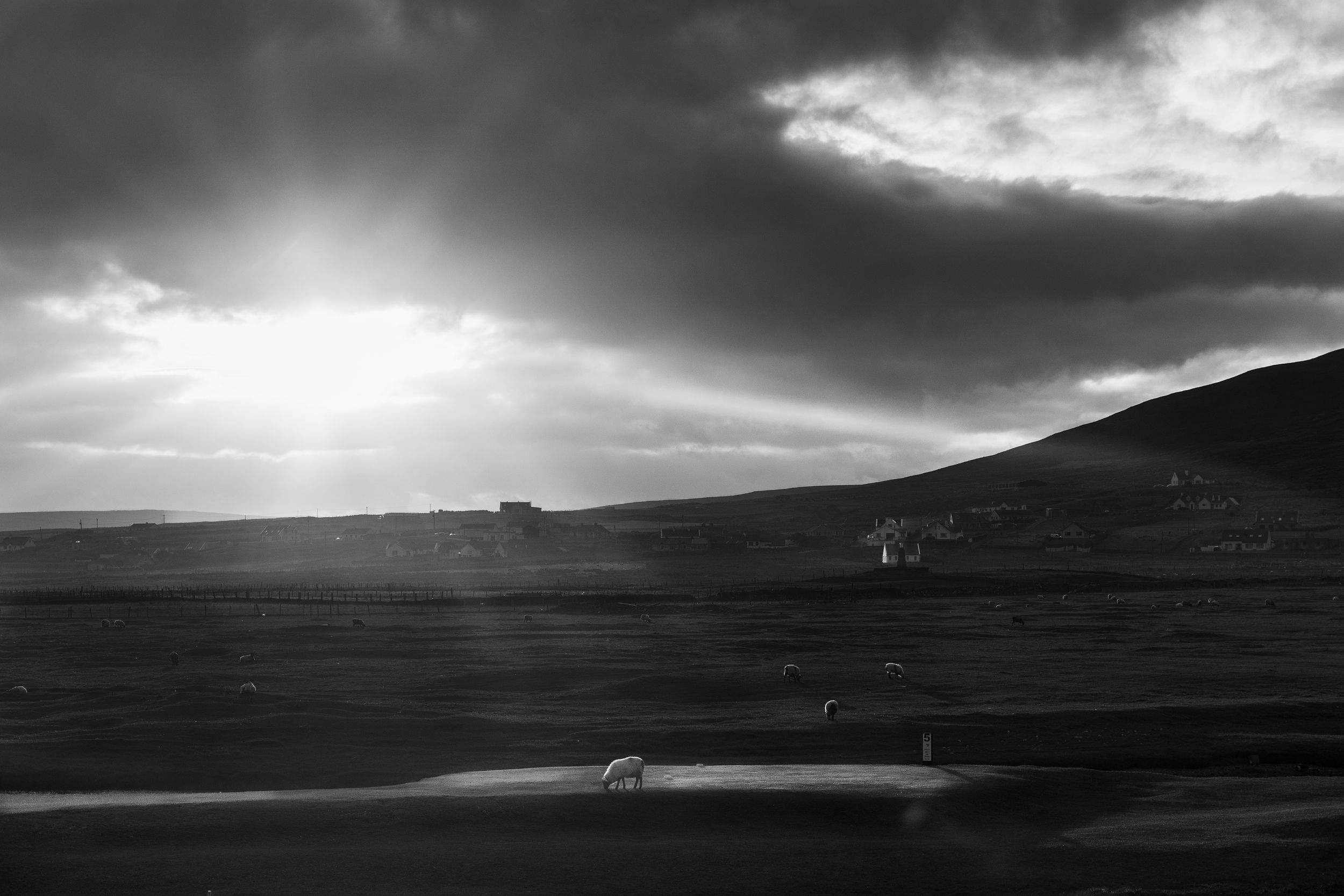 Black and white landscape photo of rolling hills under dramatic sunlit cloudy sky at dusk.