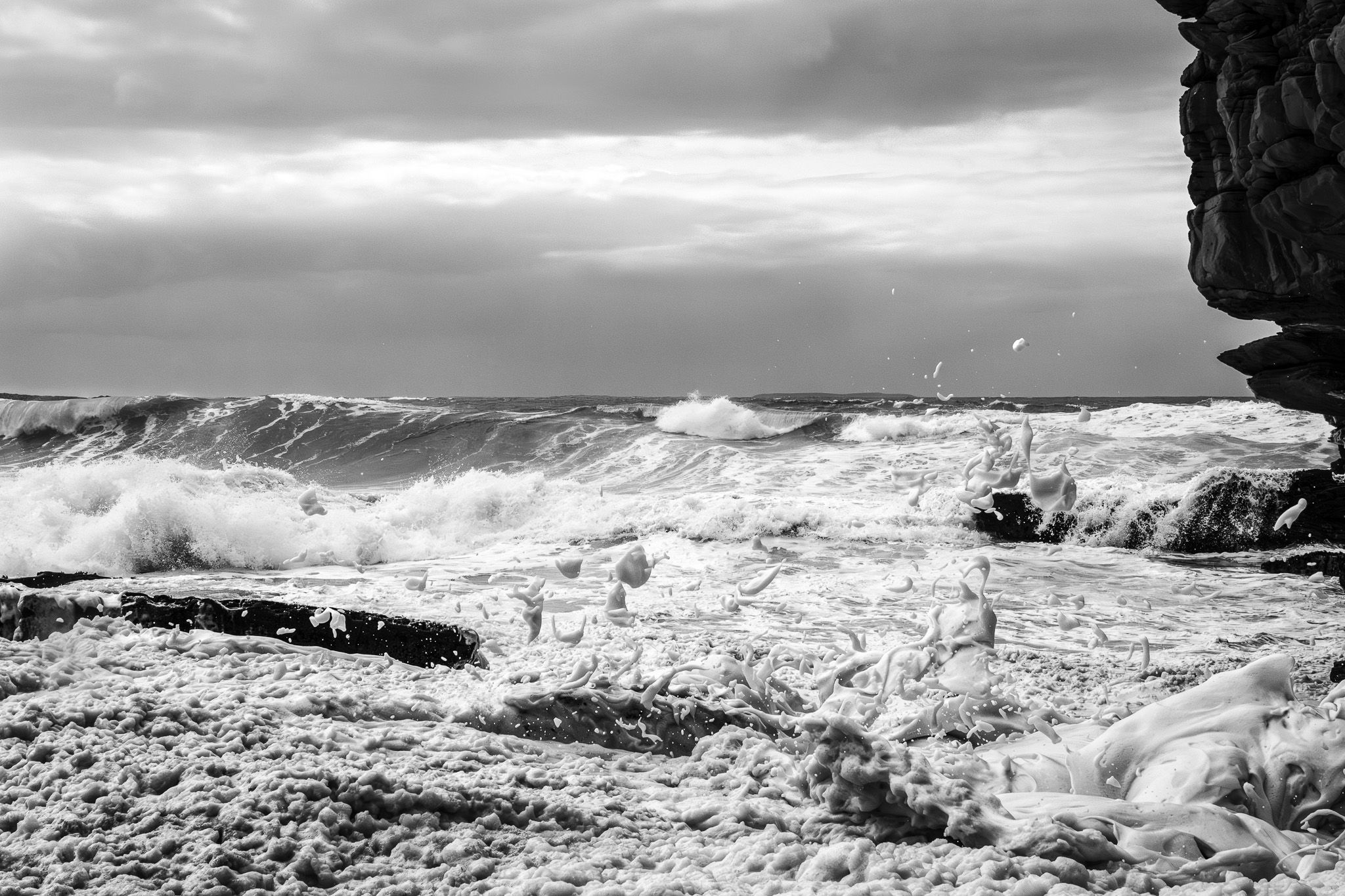 Black and white photo of stormy ocean waves crashing on rocky shore under dramatic cloudy sky.