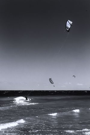 Black and white photo of kite surfers riding ocean waves with kites soaring against a dramatic sky.