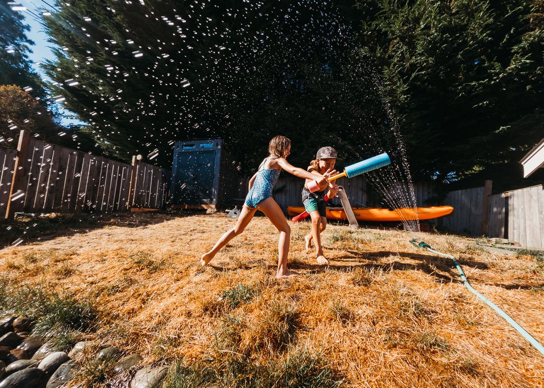 Two children playing in backyard sprinkler on sunny summer day, running through water spray on dry grass.
