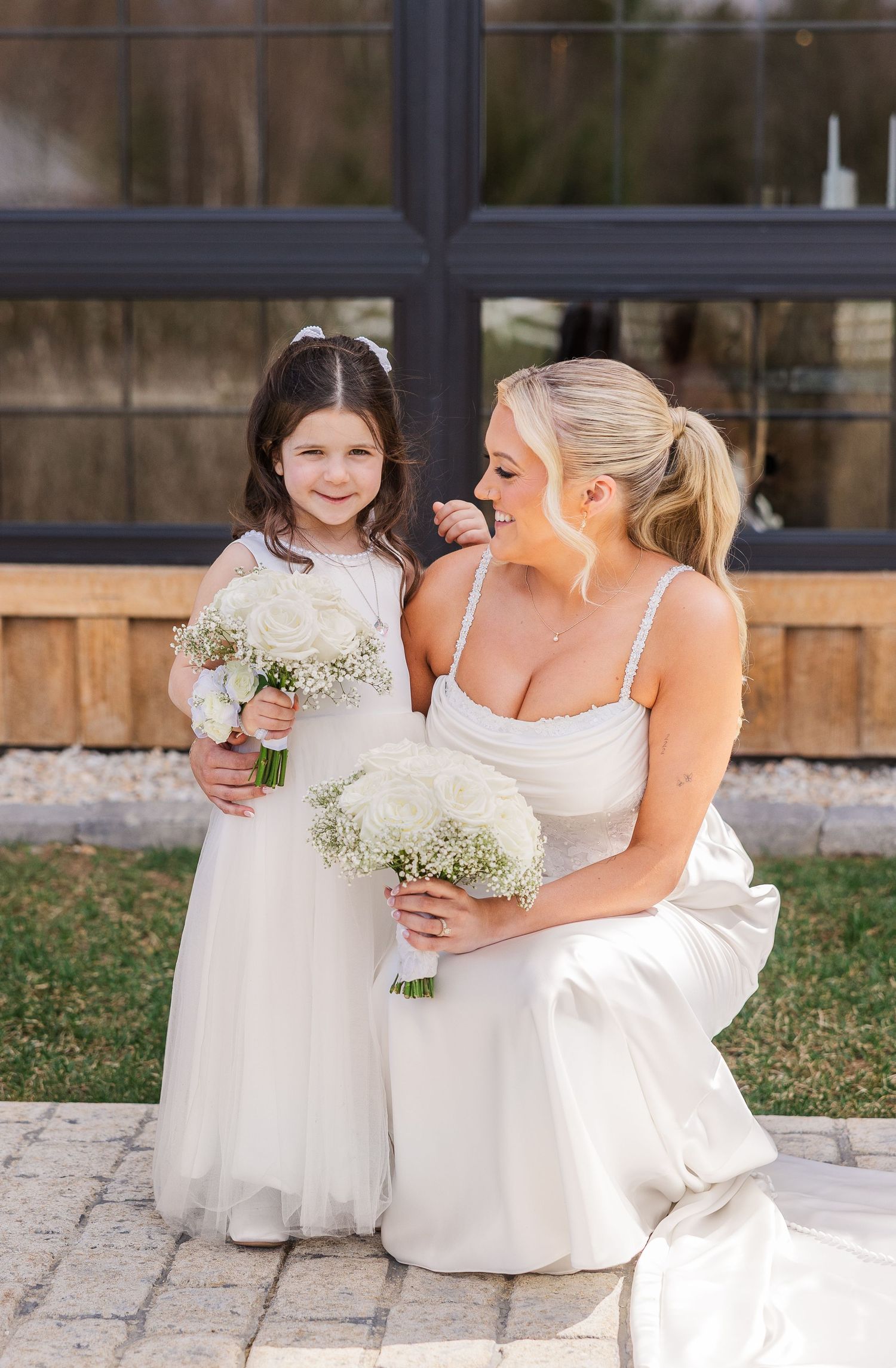 Bride crouching beside flower girl, both holding white bouquets, wearing white dresses outdoors at a wedding venue.