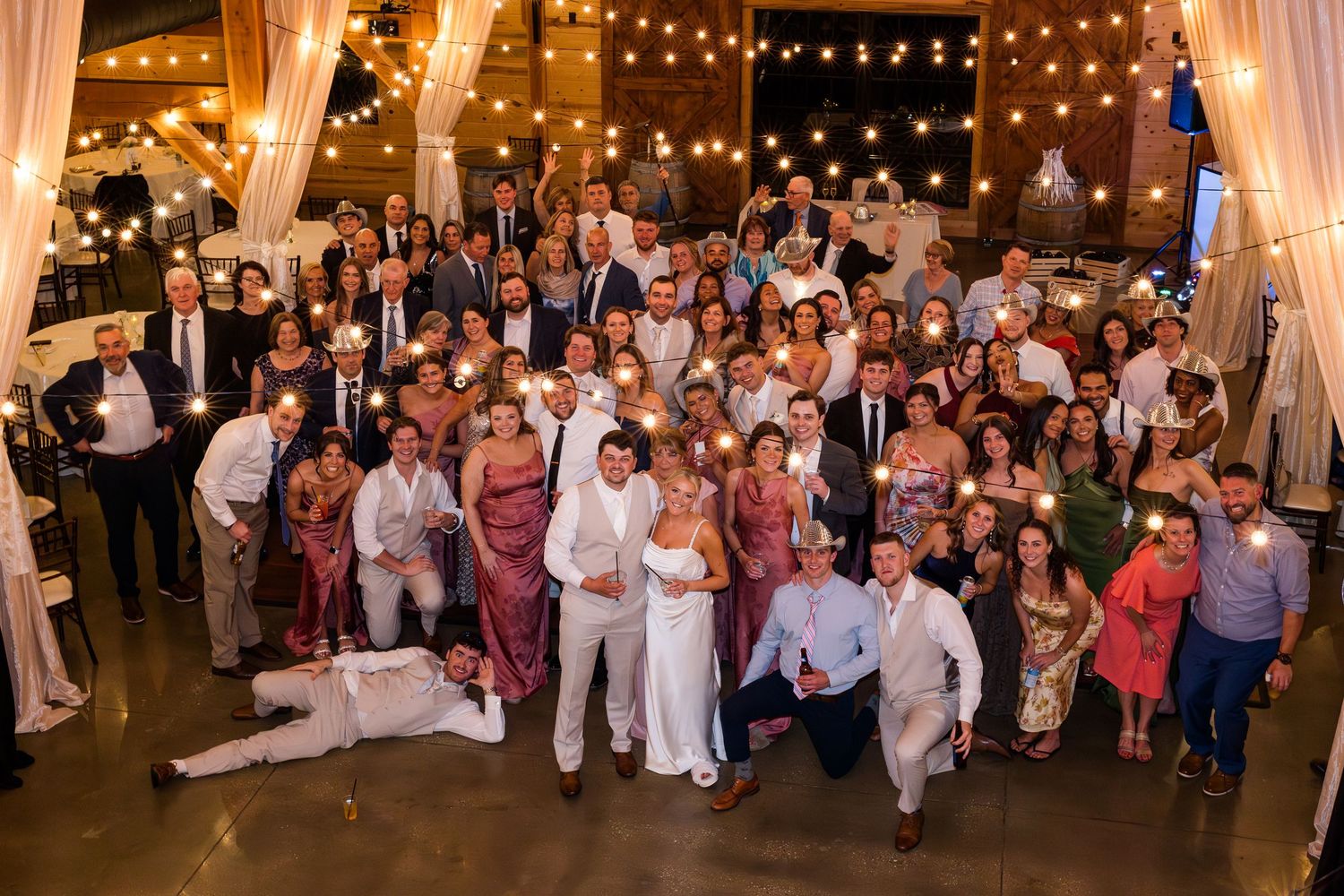 Large wedding reception group photo with bride and groom surrounded by guests holding sparklers in a warmly lit venue.