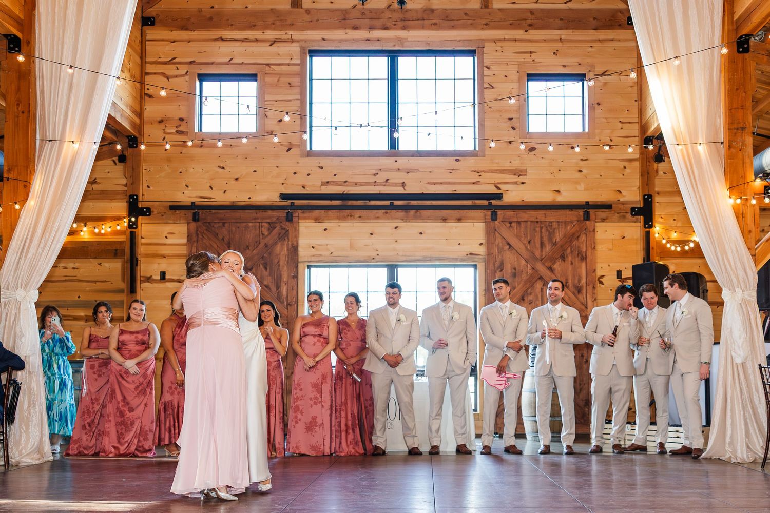 Rustic barn wedding ceremony with bridesmaids in pink dresses and groomsmen in tan suits standing before sliding wooden doors.