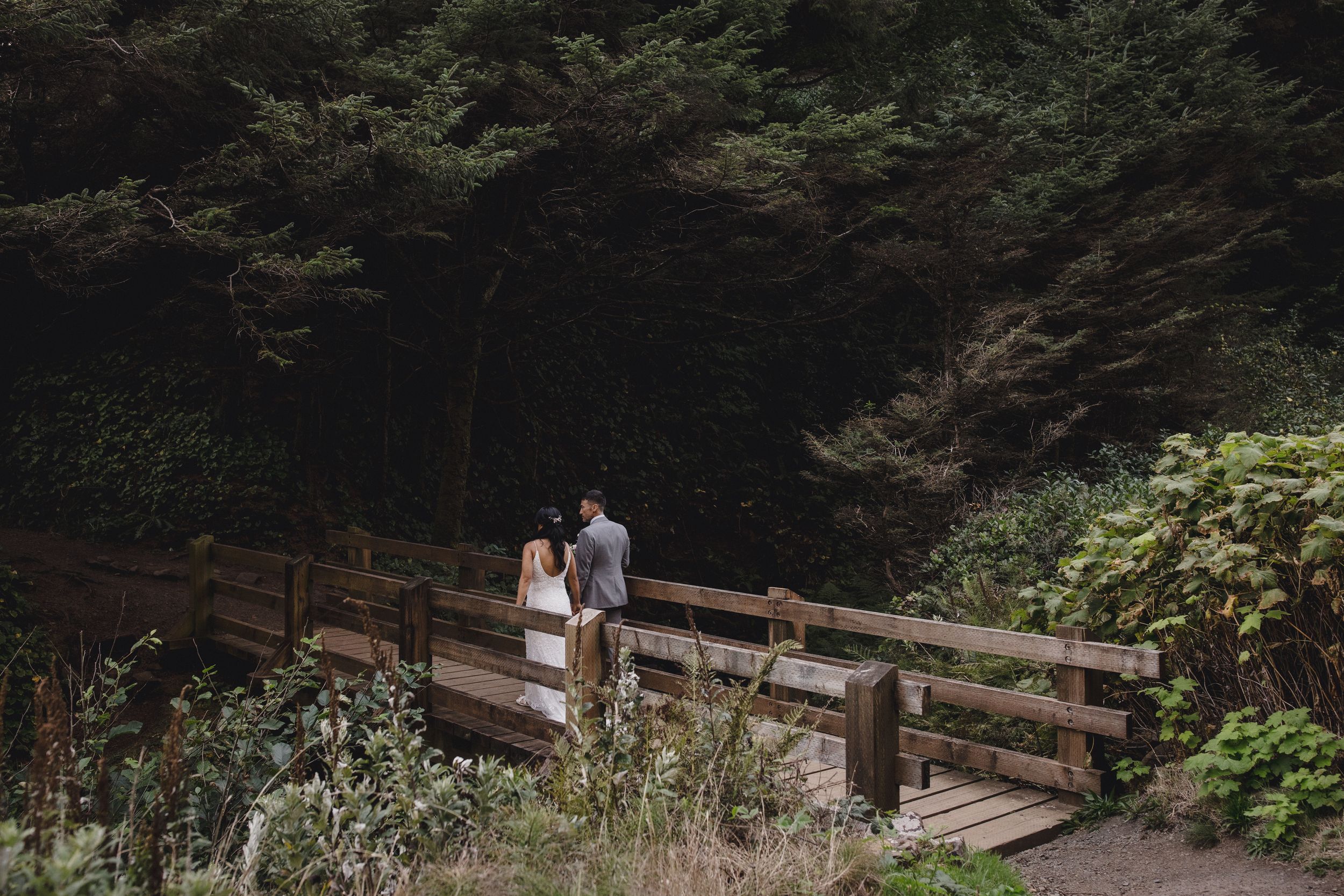 Couple standing on a wooden bridge surrounded by lush green forest trees in a romantic outdoor wedding photo.